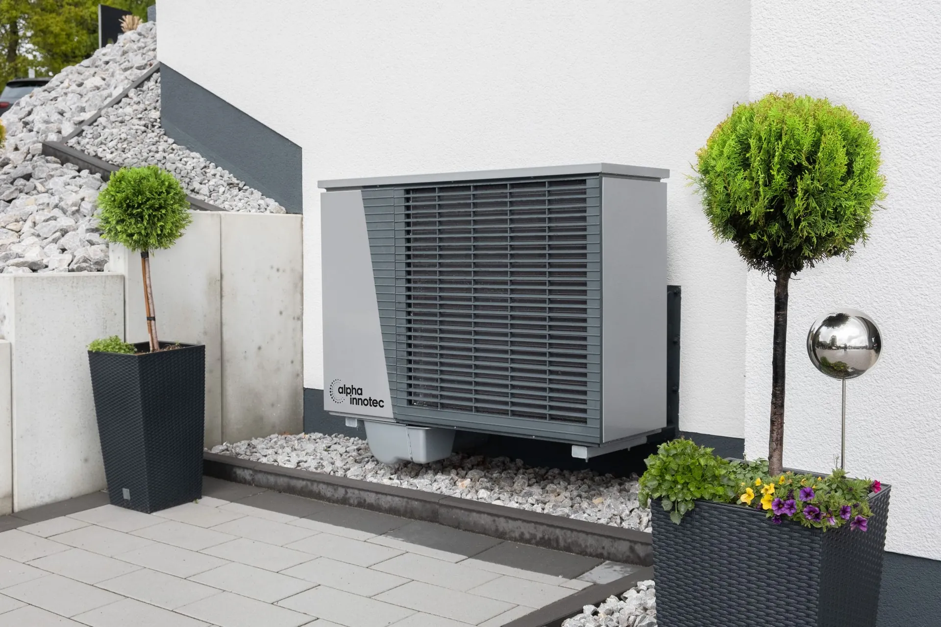 Alpha Innotec varmepumpe på hvid facade med grus og buksbomkugler i krukker